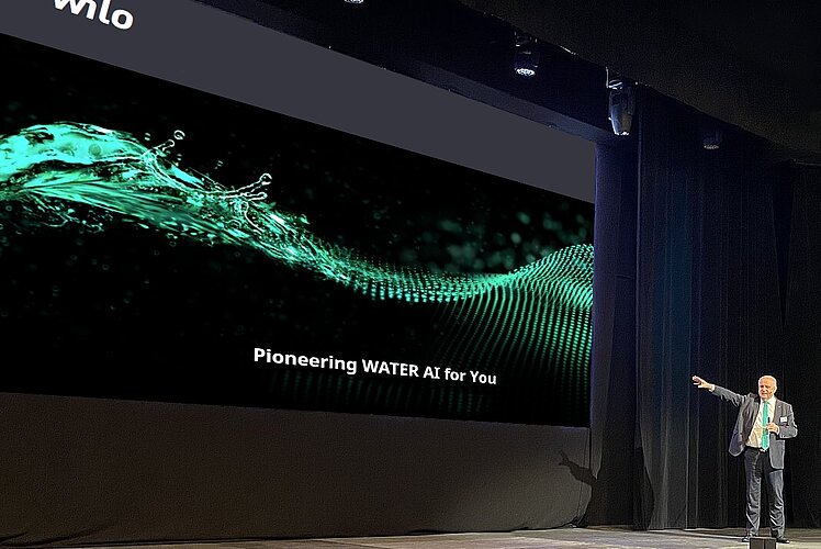 Ein Mann steht auf einer Bühne und zeigt auf ein großes Bildschirmbild, das eine grüne Wassergrafik zeigt. Darunter steht der Text "Pioneering WATER AI for You". Die Bühne ist dunkler, und die Lichtverhältnisse betonen die Präsentation.