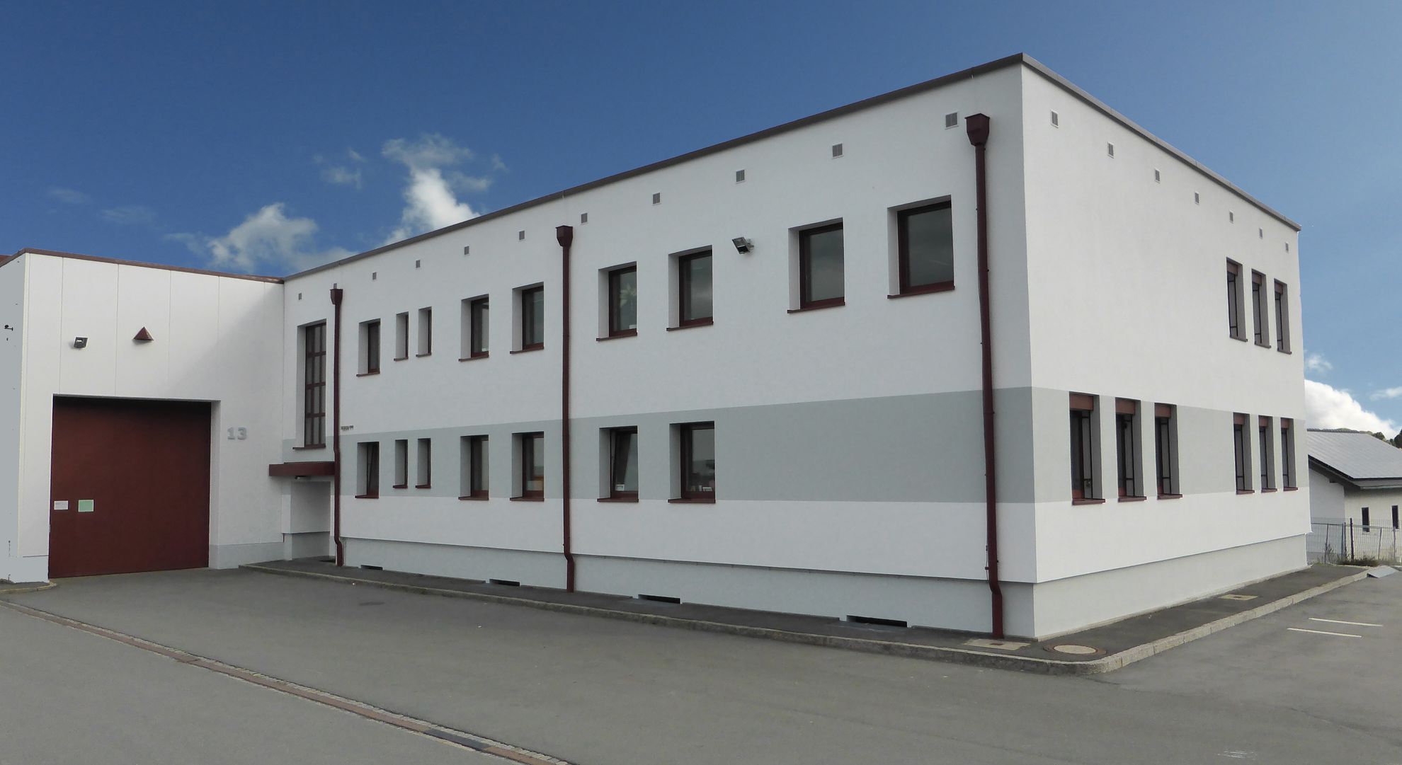 Ein modernes, zweigeschossiges Gebäude mit weißer Fassade und roten Fensterrahmen. Es hat mehrere Fenster und ein großes Eingangstor. Der Himmel ist teilweise bewölkt und es gibt einen Asphaltparkplatz vor dem Gebäude.