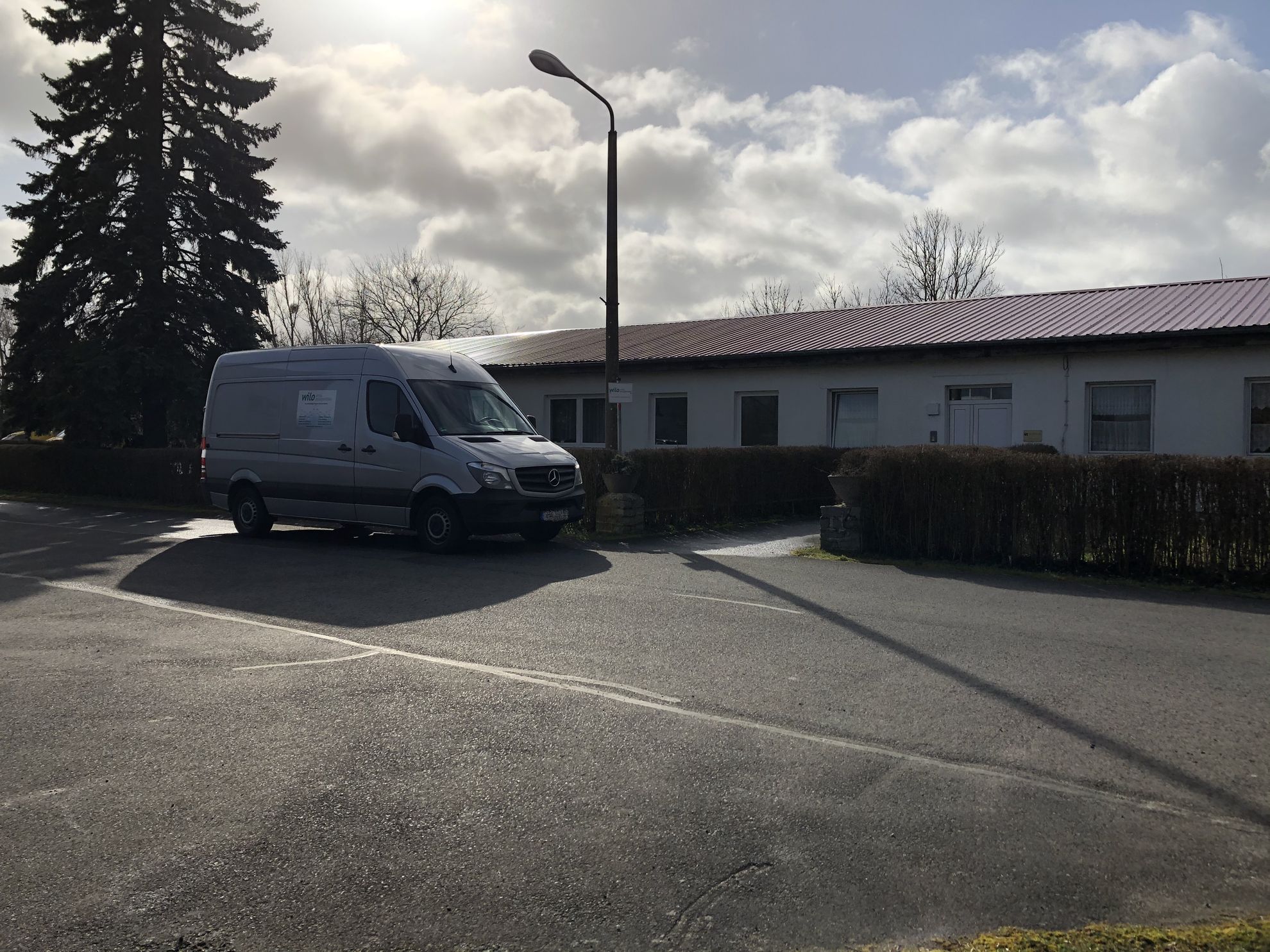 Ein weißer Lieferwagen steht auf einem Parkplatz vor einem flachen Gebäude mit Fenster. Im Hintergrund sind einige Bäume sichtbar, und der Himmel ist bewölkt, was auf wechselhaftes Wetter hindeutet.