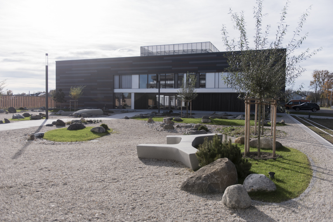Moderne Gebäude mit großen Fenstern und einem stilvollen, schwarzen Äußeren. Im Vordergrund befindet sich ein gestalteter Garten mit Kies, Steinen und Bäumen. Grüne Rasenflächen sind integriert, und eine Sitzbank ist sichtbar.
